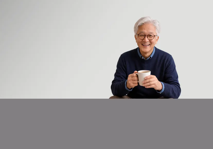 A silver-haired older person in round glasses and a dark blue jumper holds a mug in both hands, smiling happily